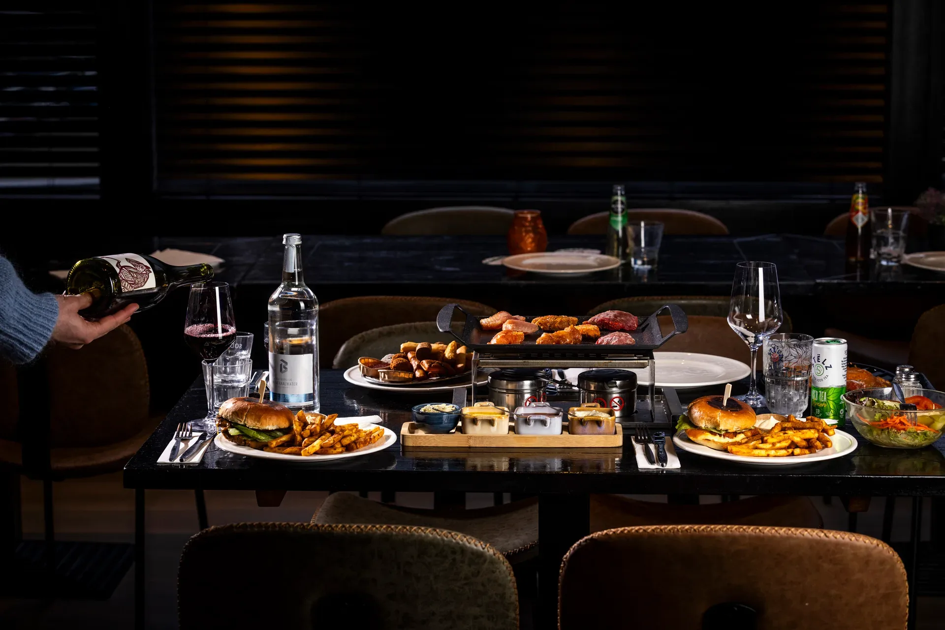 Gourmetten met verschillende vleesstukken op tafel, flessen wijn en water en borden met hamburgers en friet bij Oscars Bowling & Pool