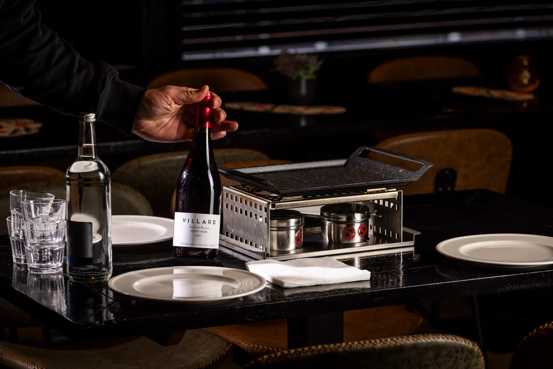 Hand die een fles rode wijn pakt aan een tafel met gourmetstel, glazen en borden bij Oscars Bowling & Pool