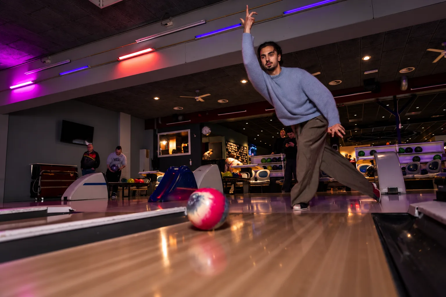 Man werpt een bowlingbal op een baan met paarse lichtaccenten bij Oscars Bowling & Pool in Zaandam, terwijl anderen op de achtergrond wachten en kijken.